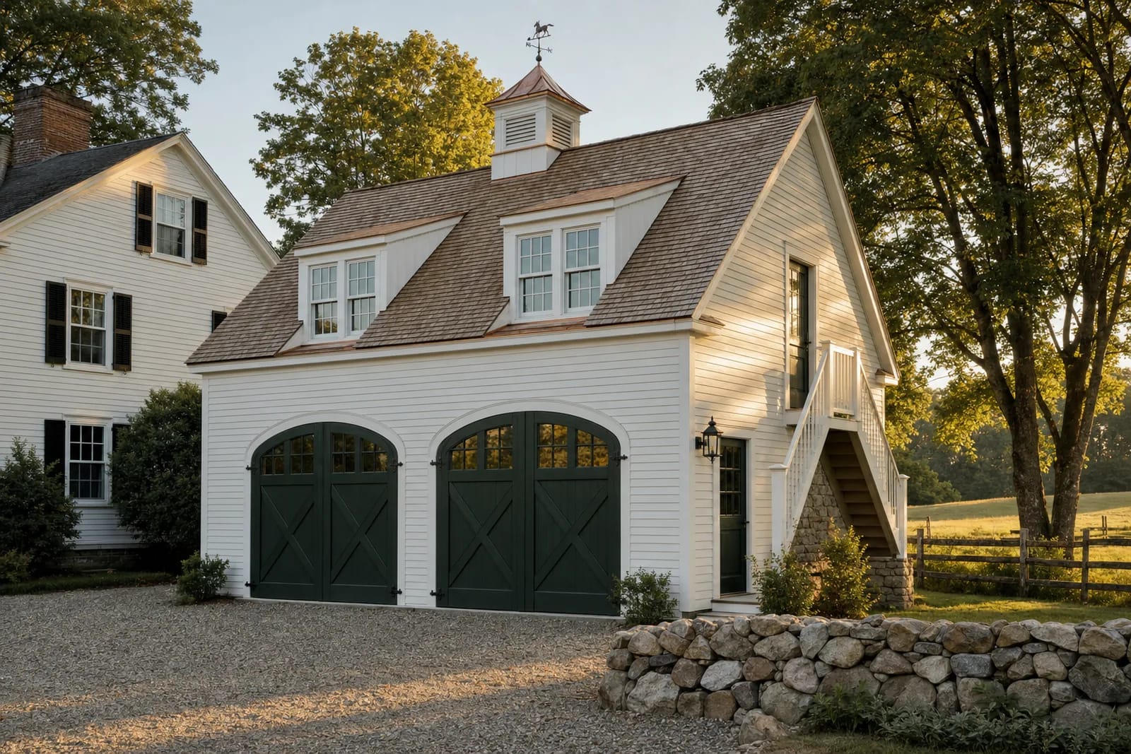 White clapboard two-bay carriage house with arched dark green carriage doors, two shed dormers, a copper-roofed cupola and an exterior stair to the upstairs apartment, on a gravel court with a fieldstone wall in late-afternoon sun