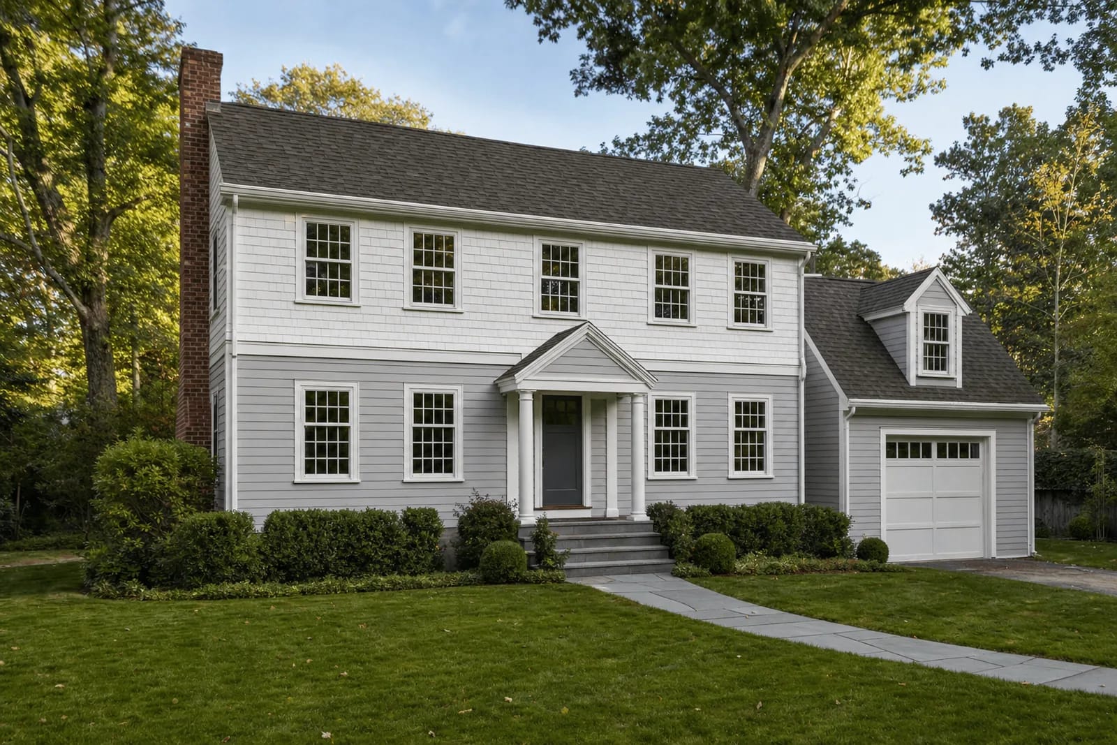 Two-story house with gray clapboards on the first floor and white shingles on the new second floor, aligned six-over-six windows, a columned entry portico and an attached garage with a dormer, on a lawn under mature oaks